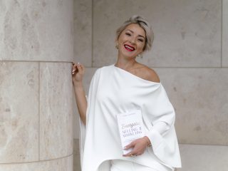 Lenka Lutonska, founder of Business Energetics®, in a camel trench coat and brown tailored outfit, outdoor editorial portrait for Brand You Magazine